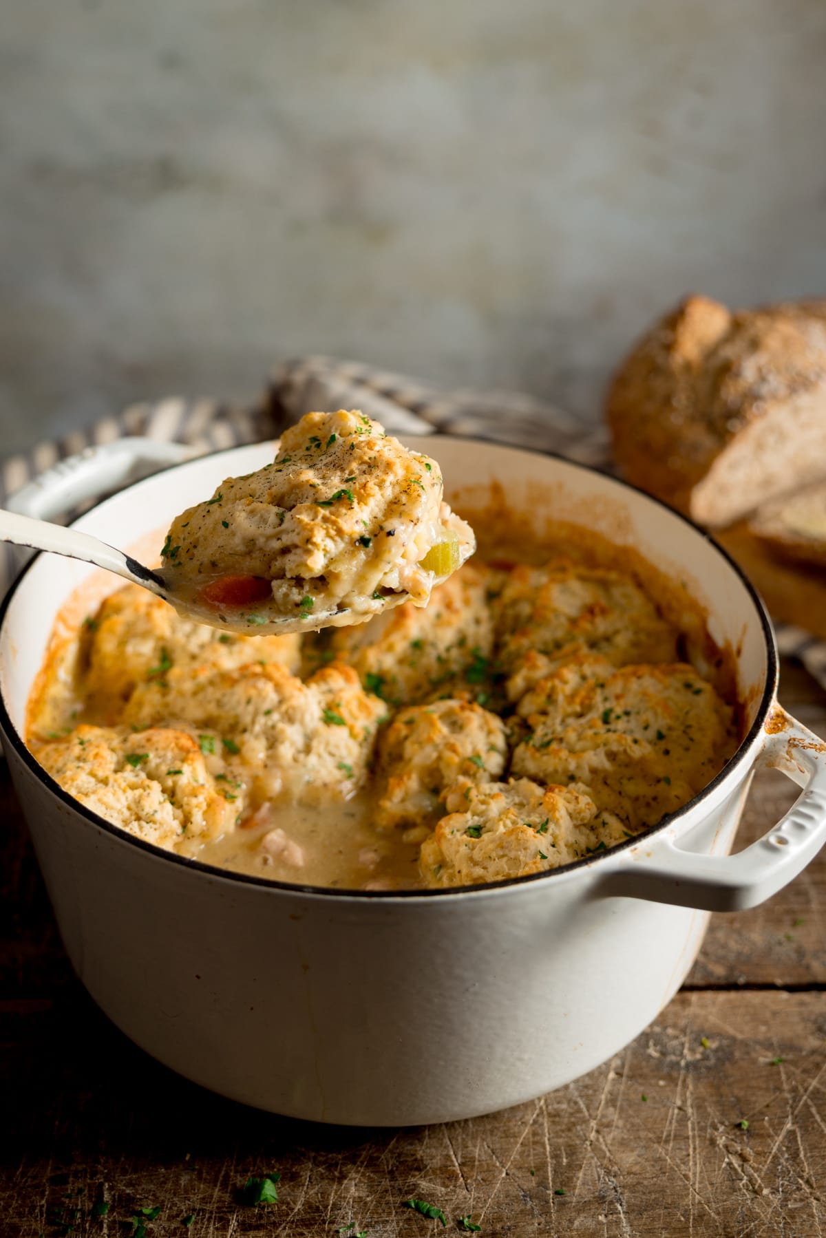 Side image of chicken stew and dumplings in a white casserole pan. A spoonful of the stew with dumpling is being lifted out of the pan. The pan is on a wooden table. There is a cut loaf of bread next to the pan.