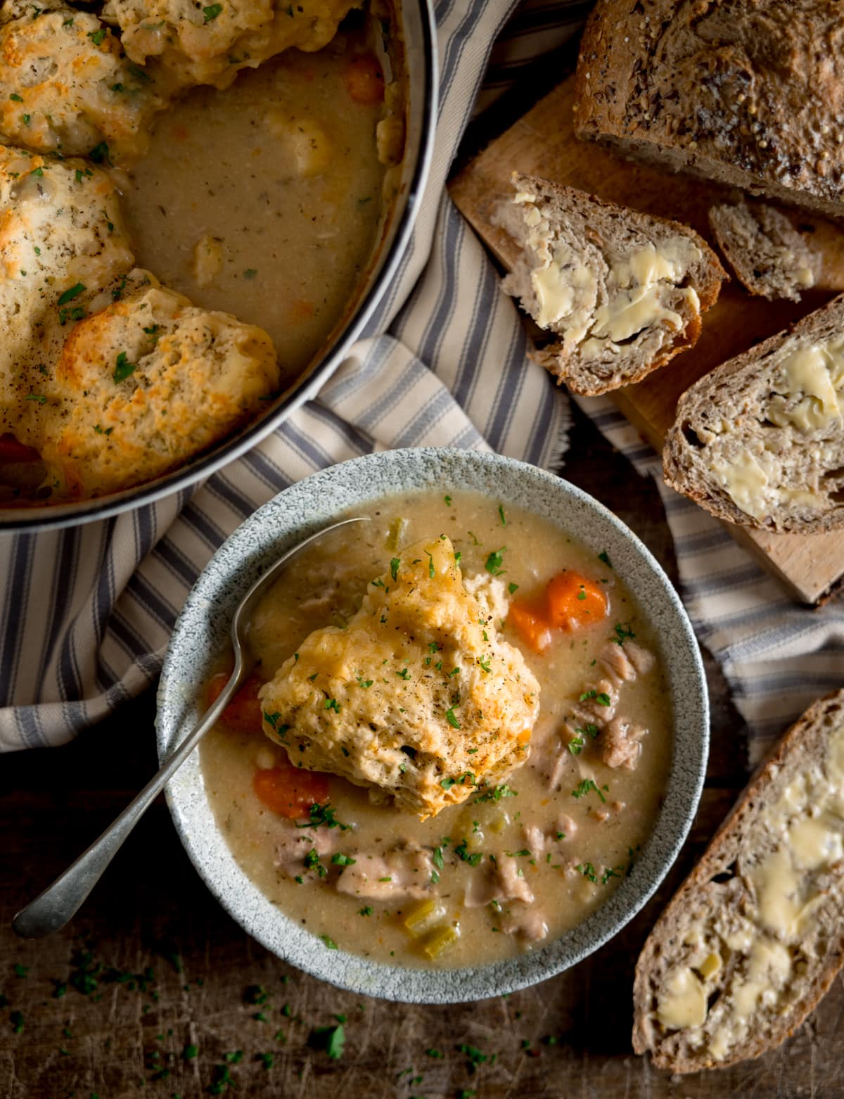 Overhead image of a serving of chicken stew and dumplings in a bowl. There is a spoon sticking out of the bowl. The bowl is on a wooden table next to a white casserole pan, filled with more stew and dumplings. There is a striped napkin and a loaf of bread with some slices cut and buttered next to the bowl.