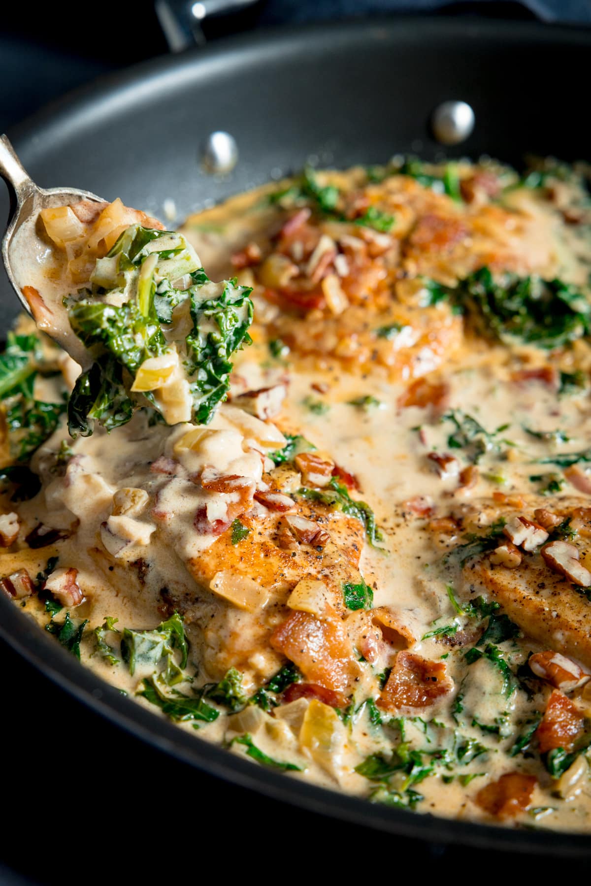 Close up picture of cream dijon sauce with kale and bacon being spooned over a cooked chicken breast in a frying pan.