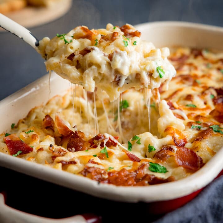 A serving spoon full of four cheese mac n cheese being lifted from a baking dish full of mac n cheese showing the stretchy cheese pull.