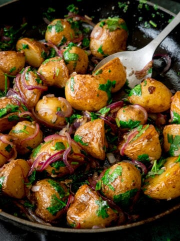 Square image of pan-fried potatoes with garlic, coriander and red onion in a black pan. There is a silver serving spoon sticking out of the pan.