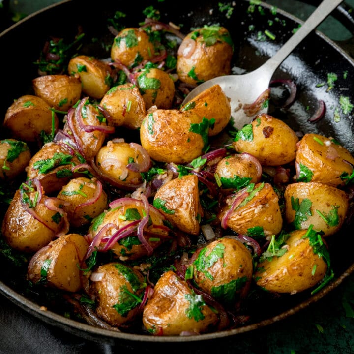 Square image of pan-fried potatoes with garlic, coriander and red onion in a black pan. There is a silver serving spoon sticking out of the pan.