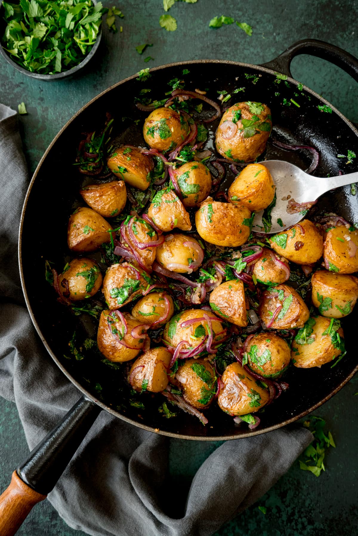 Overhead image of pan-fried potatoes with garlic, coriander and red onion in a black pan with a wooden handle. There is a silver serving spoon sticking out of the pan. The pan is on a dark green surface next to a grey napkin. There is a small bowl of chopped coriander at the top of the image.