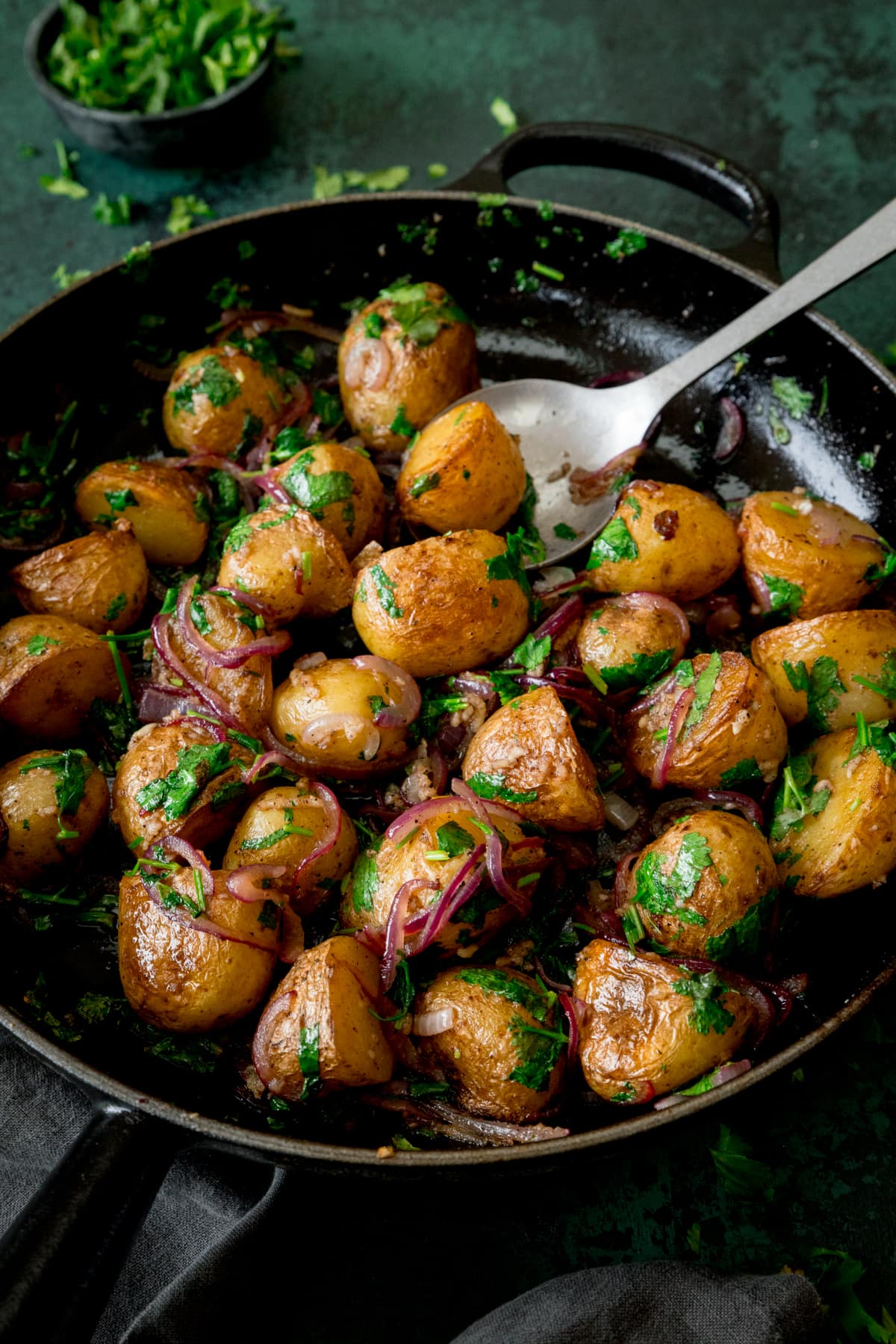 Pan-fried potatoes with garlic, coriander and red onion in a black pan. There is a silver serving spoon sticking out of the pan. The pan is on a dark green surface next to a grey napkin. There is a small bowl of chopped coriander at the top of the image.