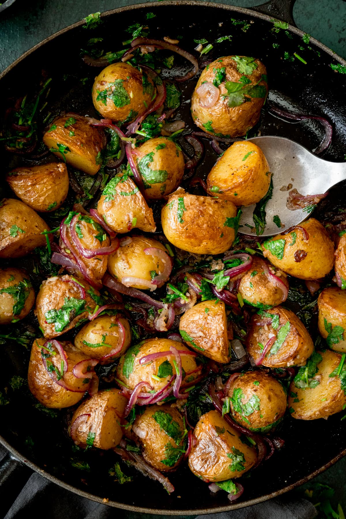 Overhead image of pan-fried potatoes with garlic, coriander and red onion in a black pan. There is a silver serving spoon sticking out of the pan.