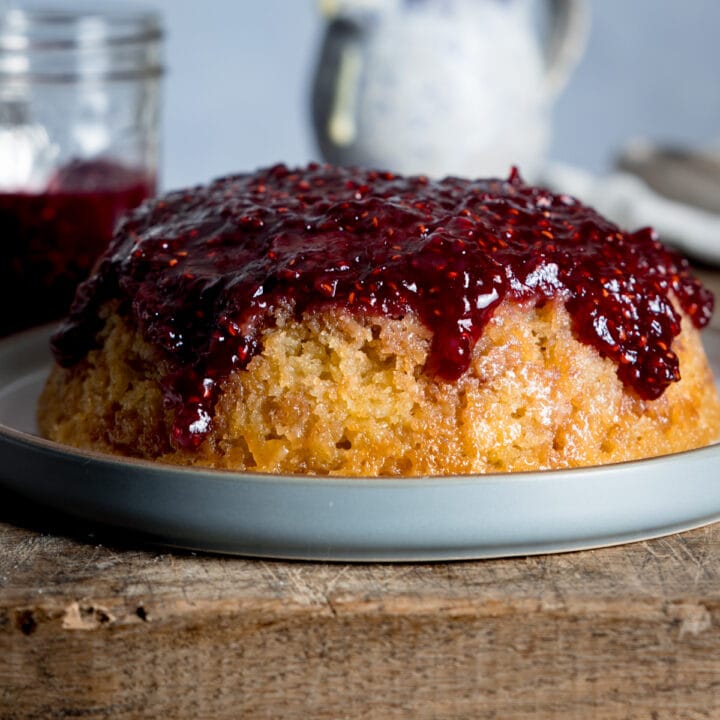a slow cooker steamed jam sponge cake on a grey plate sat on a wooden chopping board.