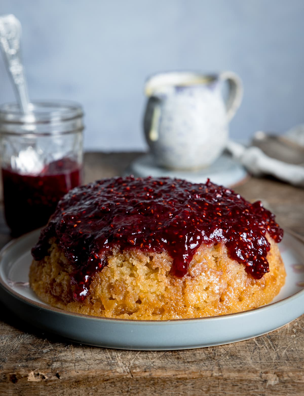 Picture of a steamed jam sponge on a grey plate thats sat on a wooden board with a jug of custard in the background.