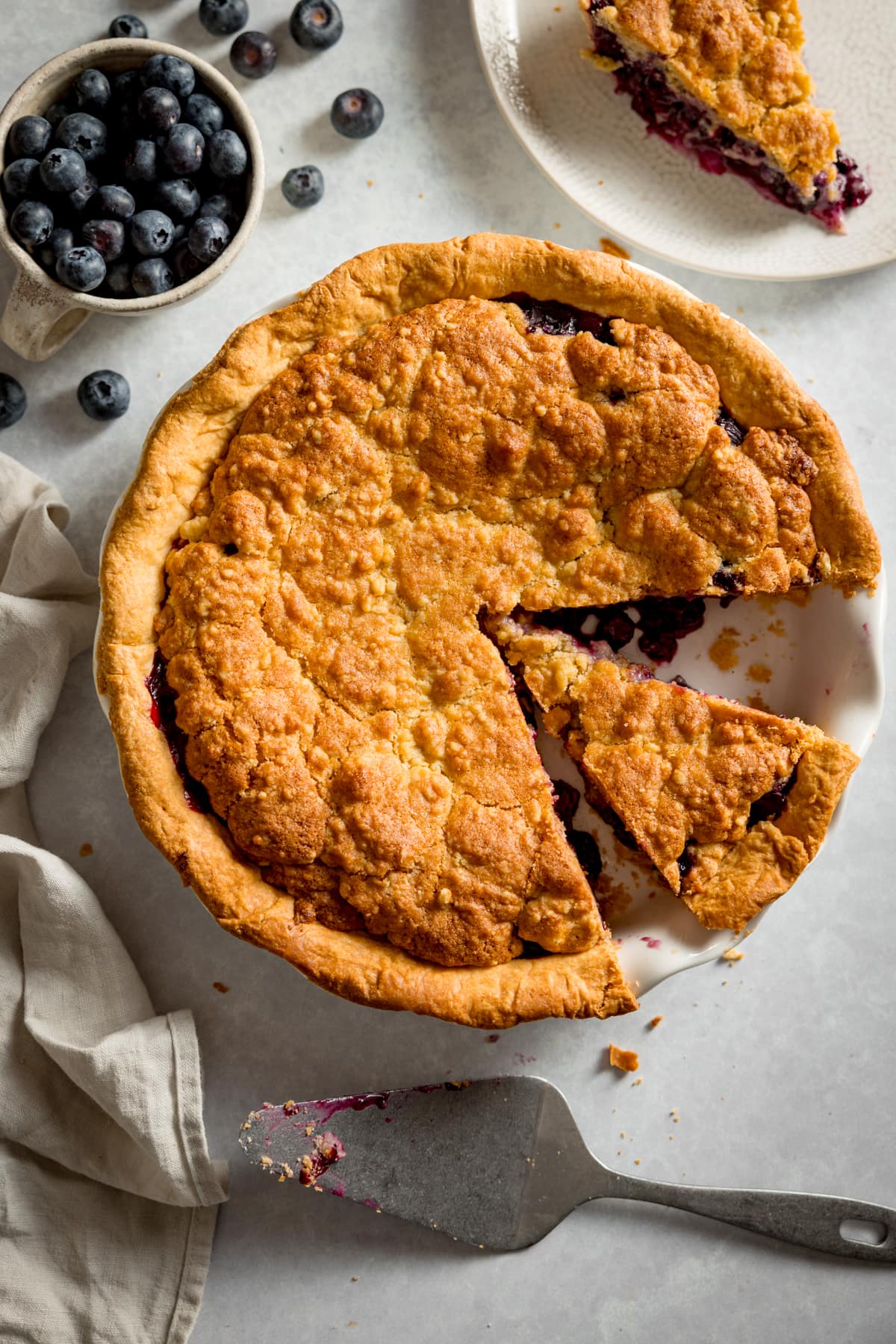 Overhead image of a blueberry crumble pie in a white pie dish with a slice removed and a further slice made, but left in the dish.
The dish is on a white background and a slice of pie is on a white plate at the top of the frame.
There is a stoneware cup filled with blueberries and blueberries scattered round at the top of the frame.
There is a silver cake slice at the bottom of the frame and a light grey napkin on the left of the pie.