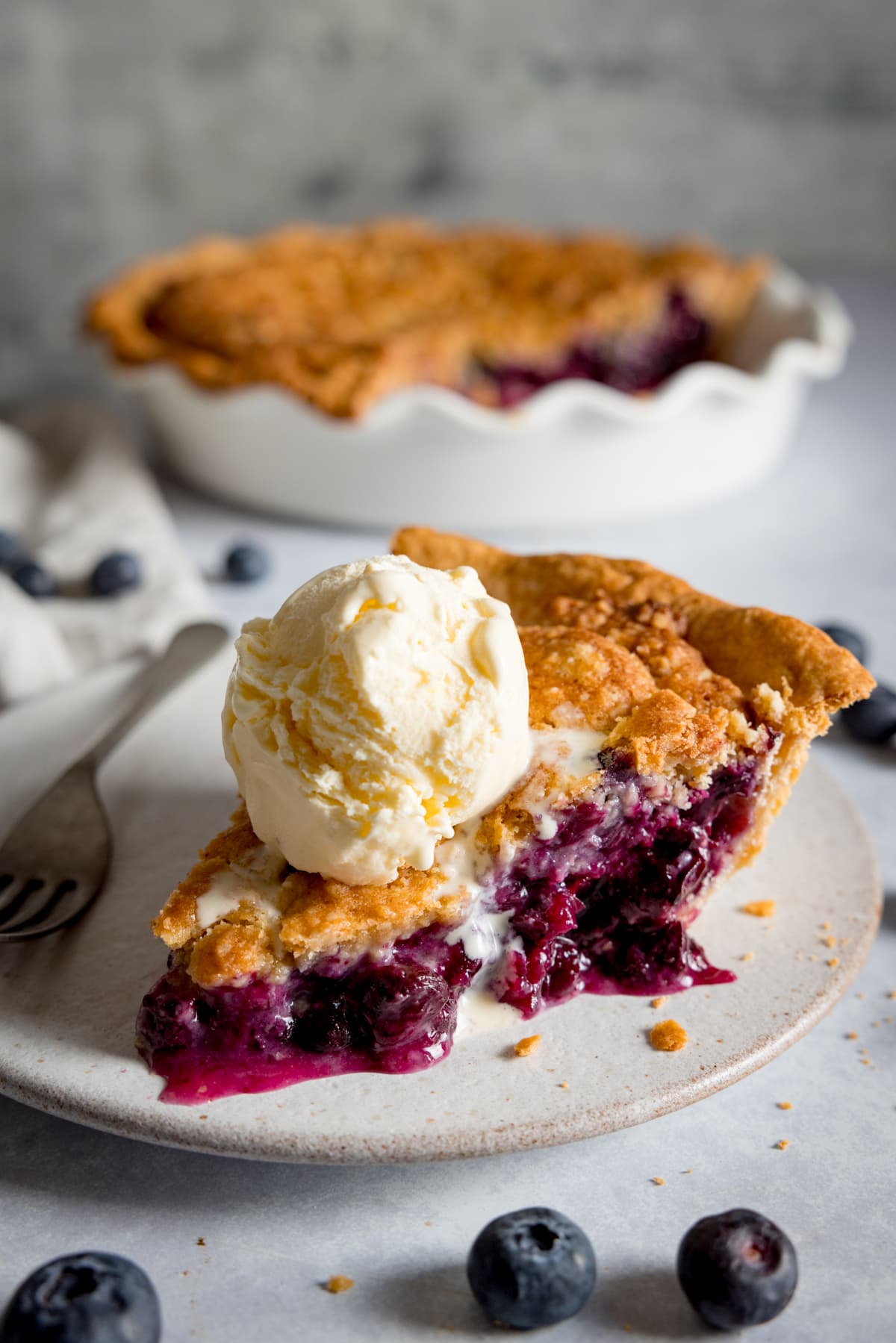 A slice of blueberry crumble pie on a white plate, topped with a scoop of vanilla ice cream.
There is a dessert fork on the plate and the plate is on a white background.
There are fresh blueberries scattered around the plate.
The rest of the pie is in the background in a white pie dish.
