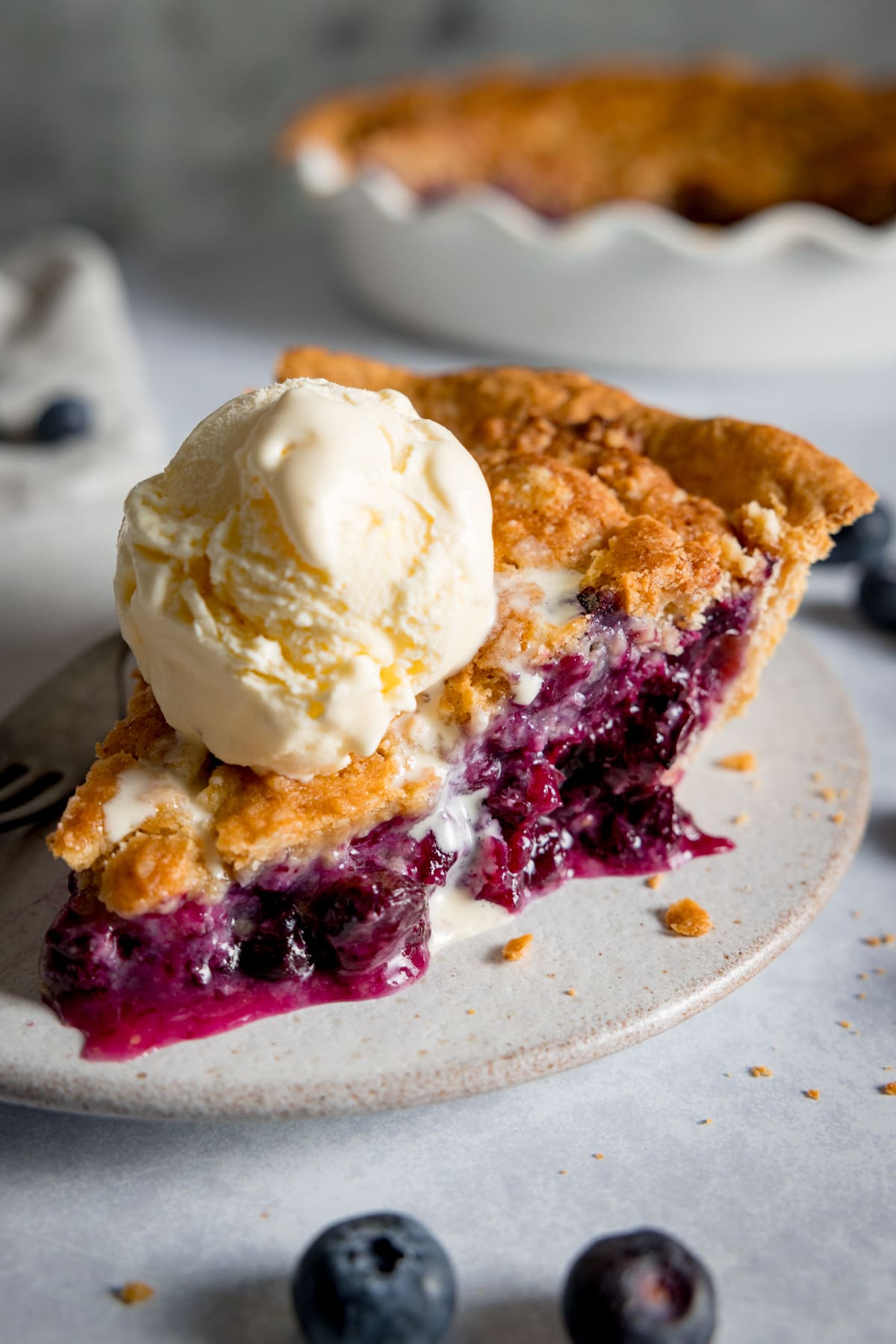 Close up of a slice blueberry crumble pie on a white plate, topped with a scoop of vanilla ice cream.
The plate is on a white background.
There are fresh blueberries scattered around the plate.
The rest of the pie is in the background in a white pie dish.