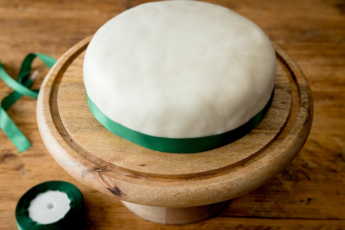 A fondant covered Christmas cake with a green ribbon on a wooden cake stand, on a wooden table. There is a roll or ribbon and a piece of ribbon next to the cake stand.