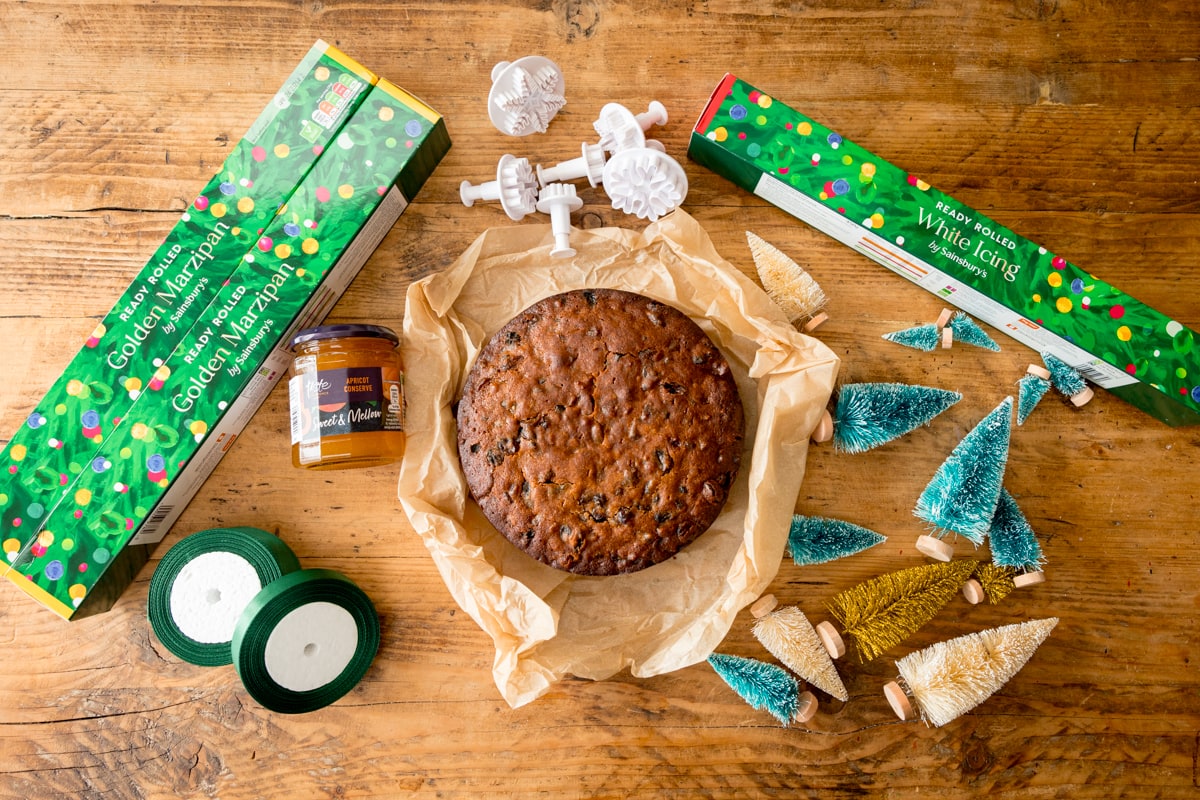 A fruitcake laying on baking parchment on a wooden table. The cake is surrounded by equipment and ingredients to decorate the cake as a Christmas cake. This includes rolls of marzipan, fondant icing, rolls of green ribbon, Apricot jam, snowflake fondant cutters and mini Christmas trees.