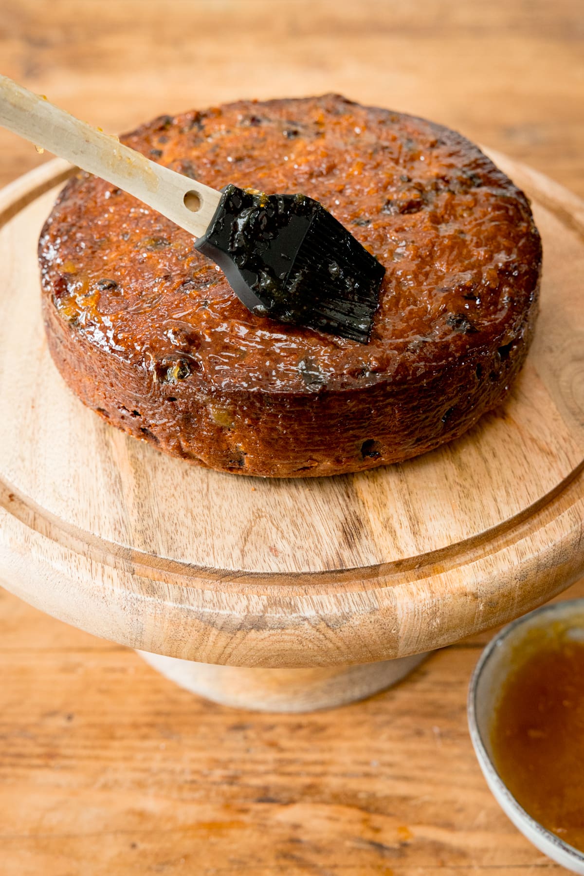 A fruitcake on a wooden cake stand, on a wooden table. The cake is being brushed with Apricot jam.