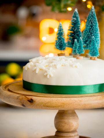 Square image showing a simple decorated Christmas cake - with a green ribbon surrounding the cake, white fondant snow flakes and mini Christmas trees on top. The cake is on a wooden cake stand on a kitchen surface with glowy lights and a bowl of fruit in the background.