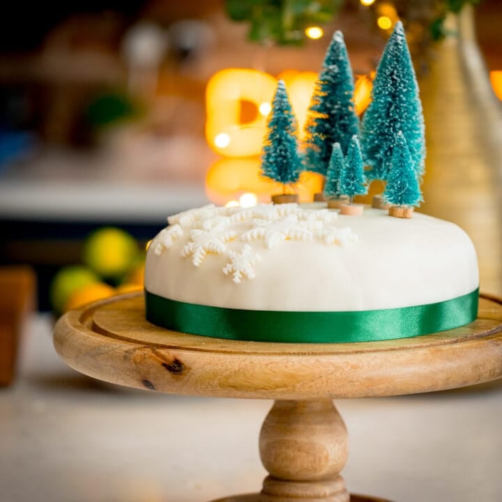 Square image showing a simple decorated Christmas cake - with a green ribbon surrounding the cake, white fondant snow flakes and mini Christmas trees on top. The cake is on a wooden cake stand on a kitchen surface with glowy lights and a bowl of fruit in the background.