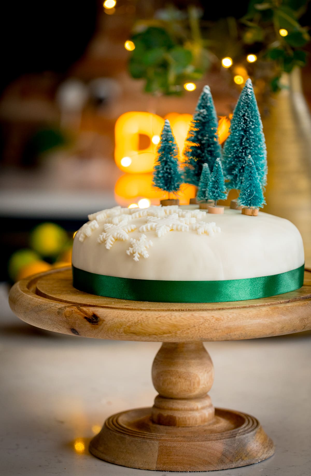 Tall side-on image showing a simple decorated Christmas cake - with a green ribbon surrounding the cake, white fondant snow flakes and mini Christmas trees on top. The cake is on a wooden cake stand on a kitchen surface with glowy lights and a bowl of fruit in the background.