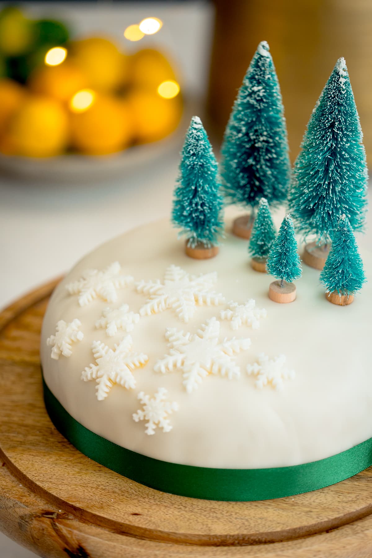 A close-up image of a simple decorated Christmas cake - with a green ribbon surrounding the cake, white fondant snow flakes and mini Christmas trees on top. The cake is on a wooden cake stand on a kitchen surface with glowy lights and a bowl of fruit in the background.