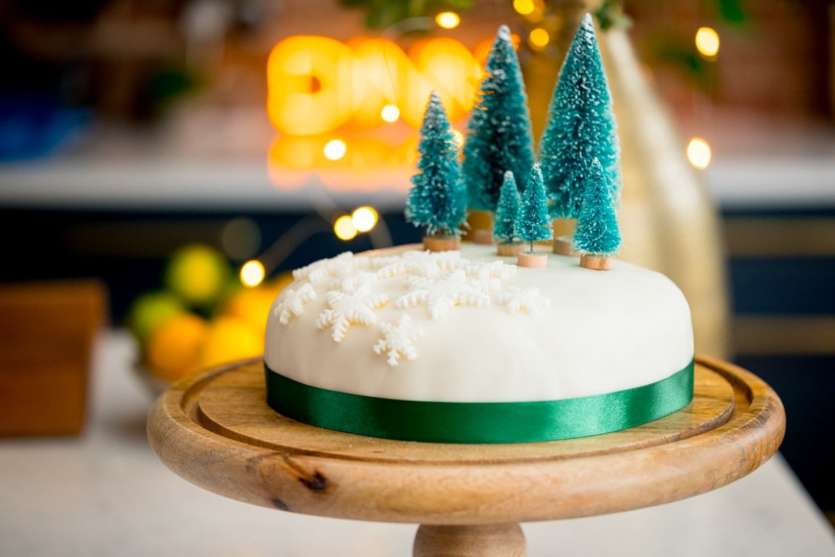 Wide image showing a simple decorated Christmas cake - with a green ribbon surrounding the cake, white fondant snow flakes and mini Christmas trees on top. The cake is on a wooden cake stand on a kitchen surface with glowy lights and a bowl of fruit in the background.