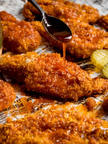 Square image of crispy baked chicken breast fillets on a baking tray with sliced pickles. The chicken is being drizzled with hot honey sauce from a black spoon.