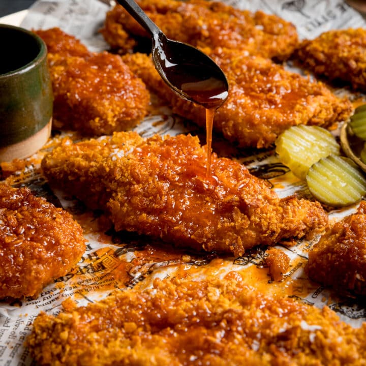 Square image of crispy baked chicken breast fillets on a baking tray with sliced pickles. The chicken is being drizzled with hot honey sauce from a black spoon.