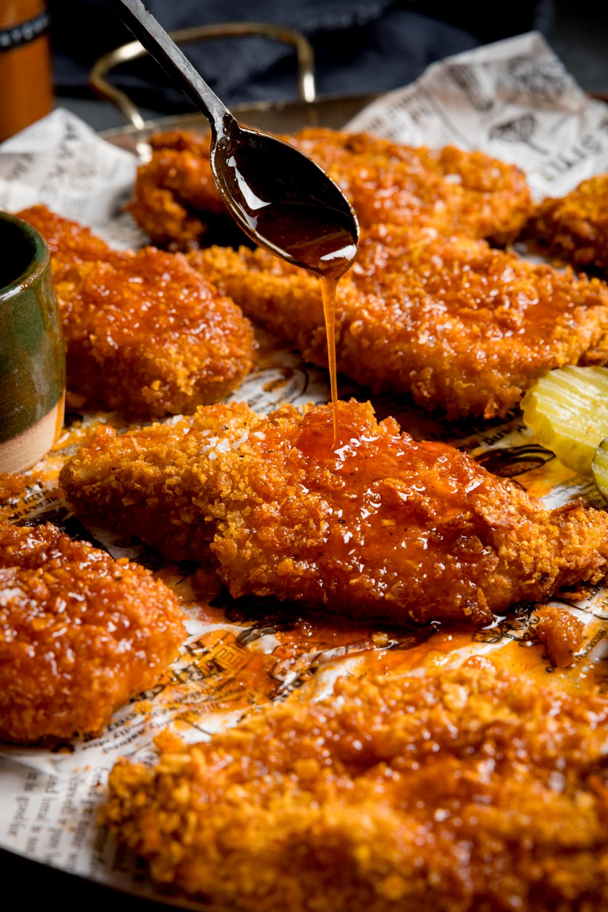 Crispy baked chicken breast fillets on a baking tray with sliced pickles. The chicken is being drizzled with hot honey sauce from a black spoon.