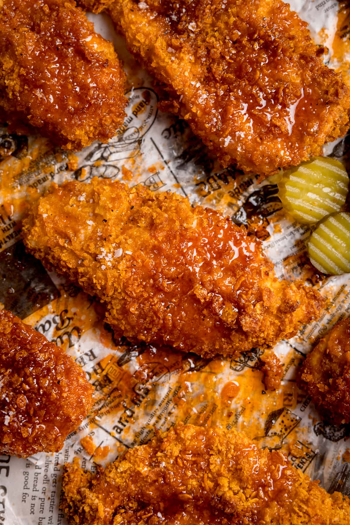 Overhead close-up image of crispy baked chicken breast fillets on a baking tray with sliced pickles. The chicken has been drizzled with hot honey sauce.