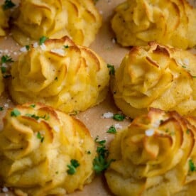 Close up square image of a tray full of piped, baked, Duchess potatoes. The tray is lined with baking parchment. The potatoes are scattered with salt and parsley.