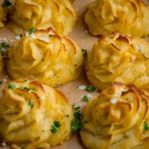 Close up square image of a tray full of piped, baked, Duchess potatoes. The tray is lined with baking parchment. The potatoes are scattered with salt and parsley.