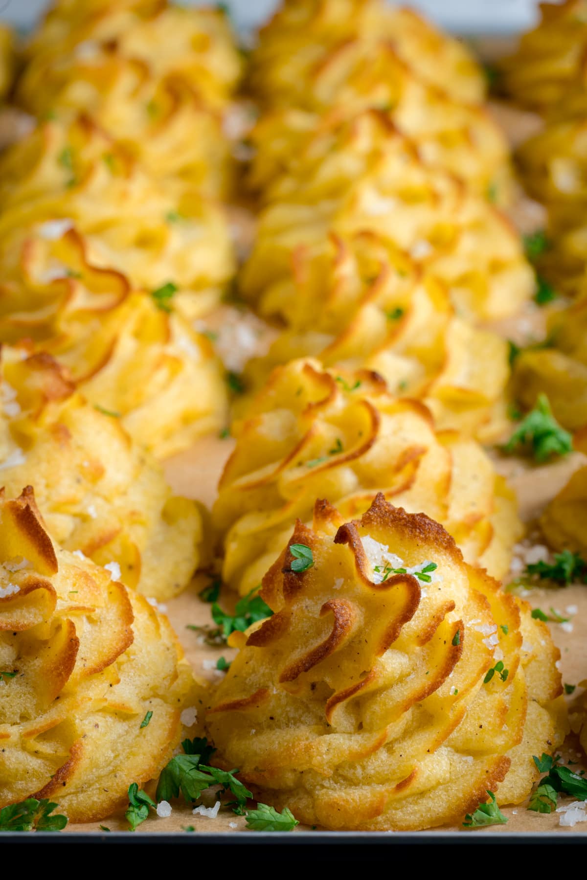 Close up image of a tray full of piped, baked, Duchess potatoes. The tray is lined with baking parchment. The potatoes are scattered with salt and parsley.