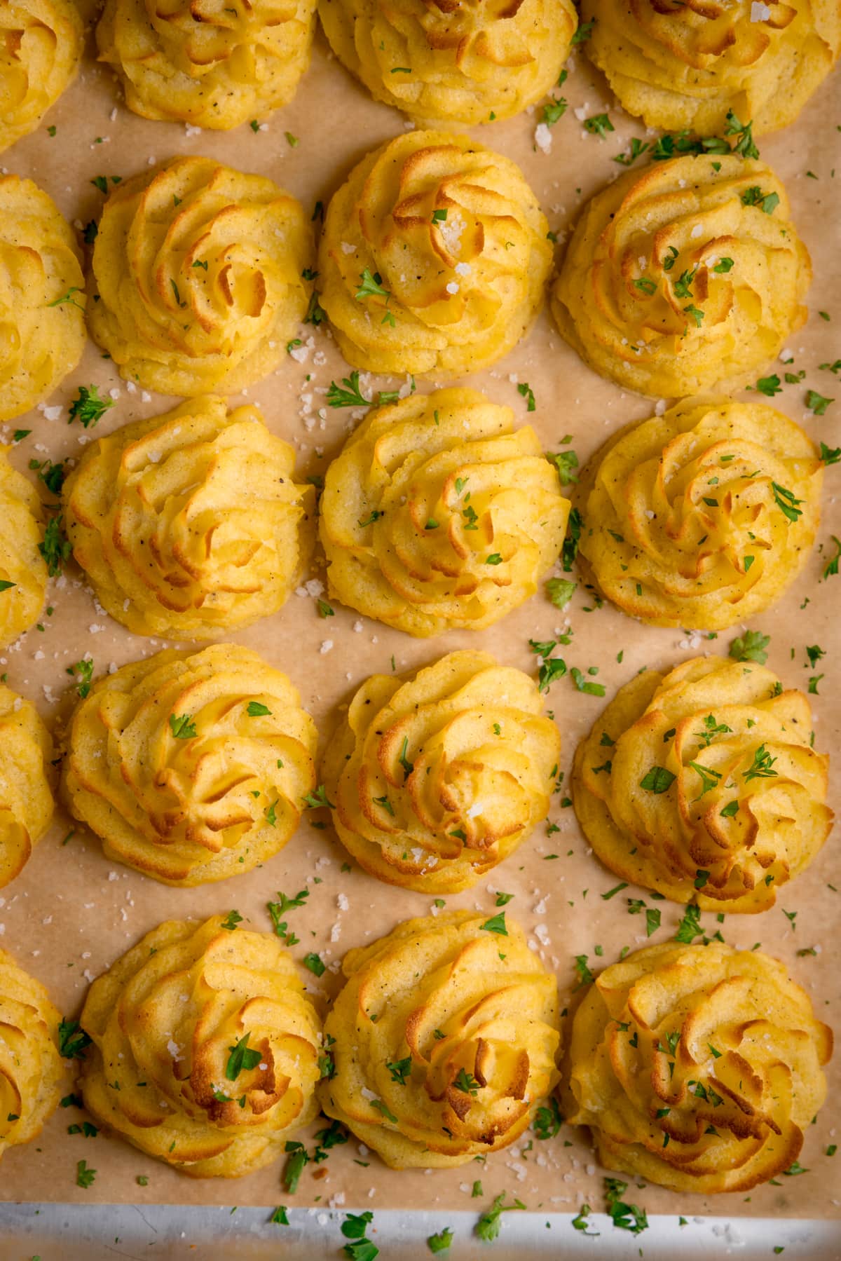 Overhead image of a tray full of piped, baked, Duchess potatoes. The tray is lined with baking parchment. The potatoes are scattered with salt and parsley.