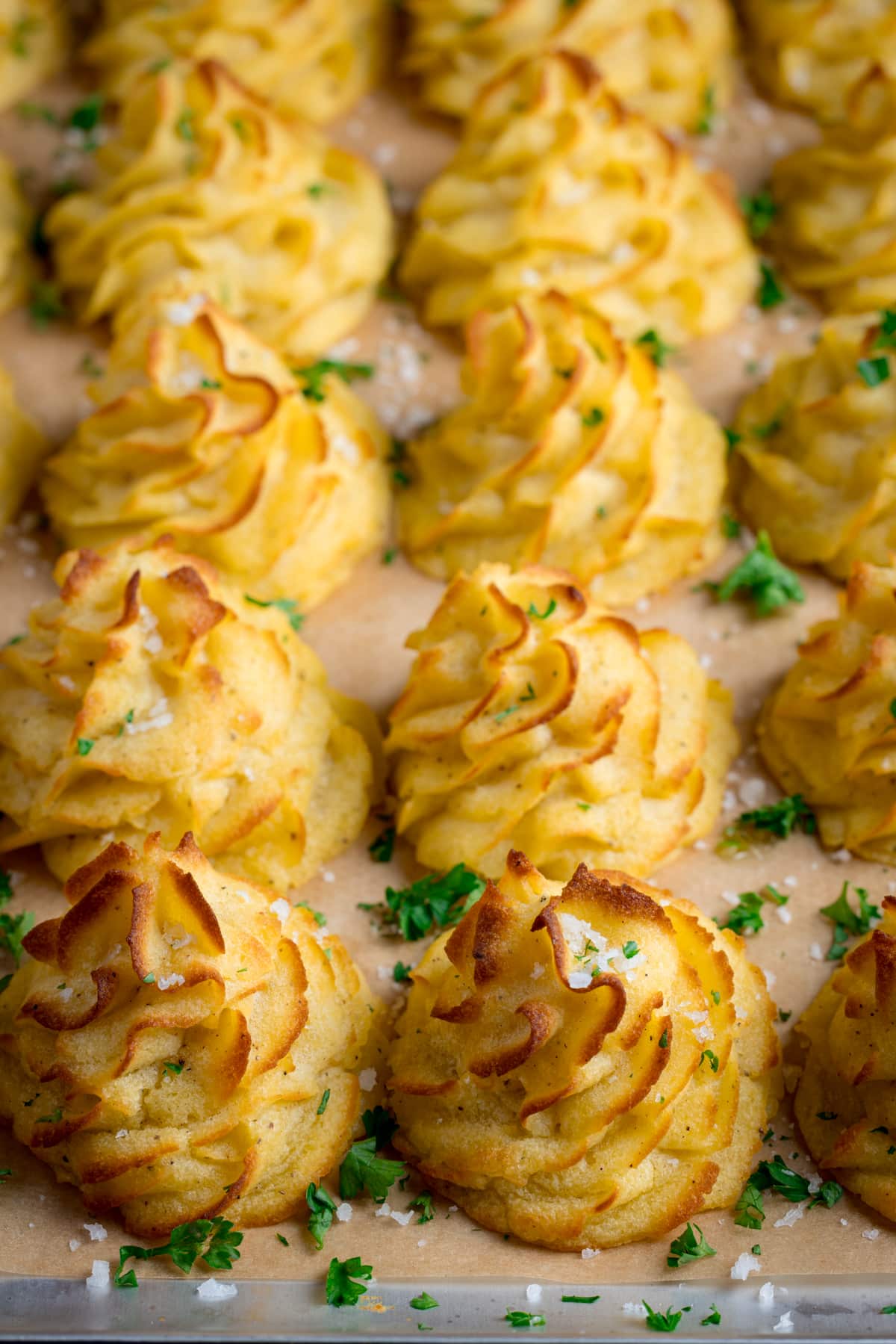 A tray full of piped, baked, Duchess potatoes. The tray is lined with baking parchment. The potatoes are scattered with salt and parsley.