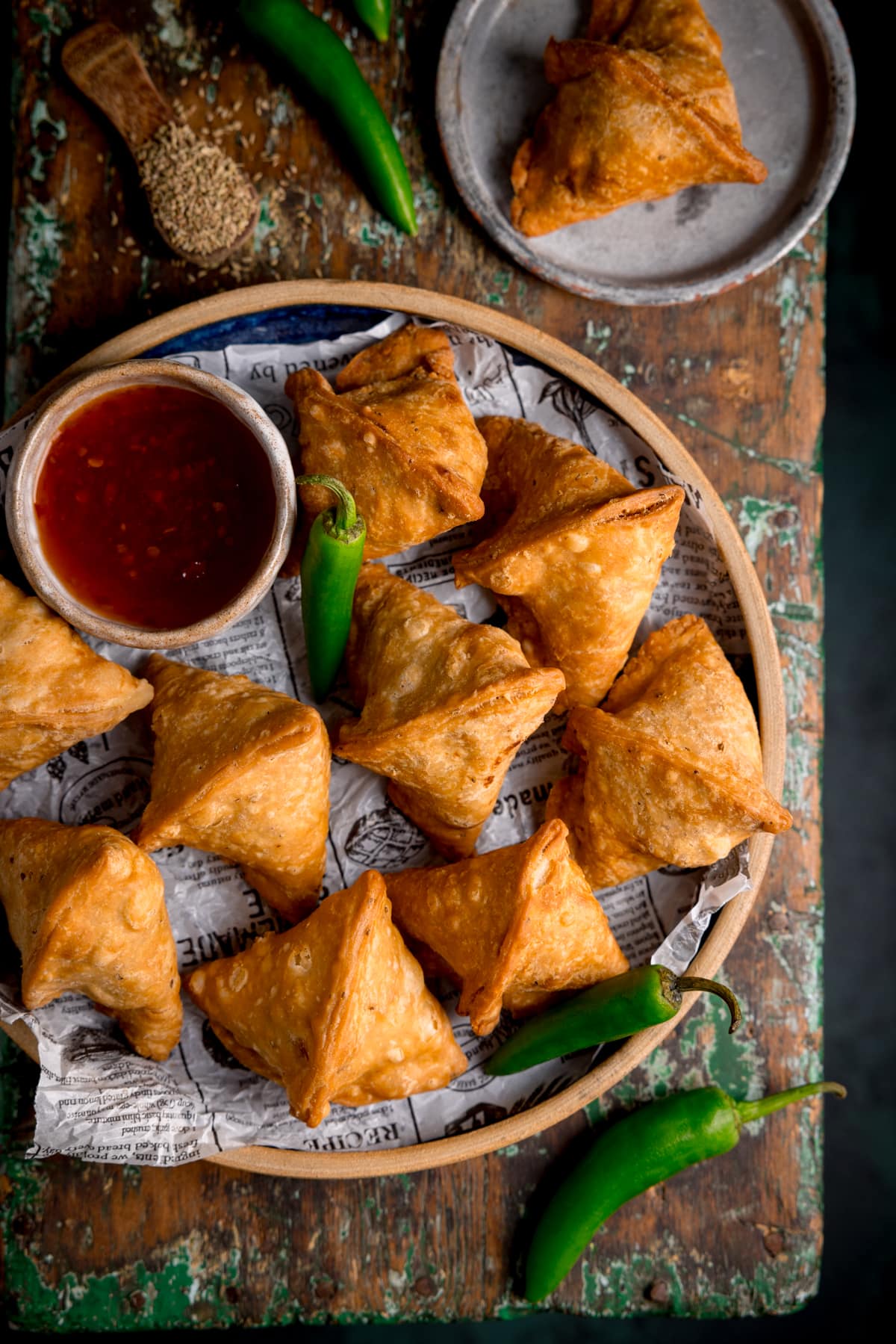 Overhead photo of a plate full of handmade Indian Samosa's with a small pot of dipping sauce all sat on a rustic wooden board.