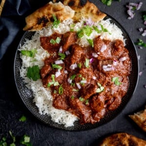 Square overhead image of slow cooked beef madras in a black bowl with rice and pieces of naan bread. There is coriander (cilantro) sprinkled on top. The bowl is on a dark surface and there are ingredients scattered around.