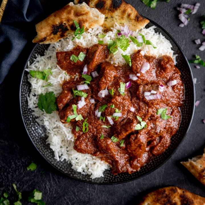 Square overhead image of slow cooked beef madras in a black bowl with rice and pieces of naan bread. There is coriander (cilantro) sprinkled on top. The bowl is on a dark surface and there are ingredients scattered around.