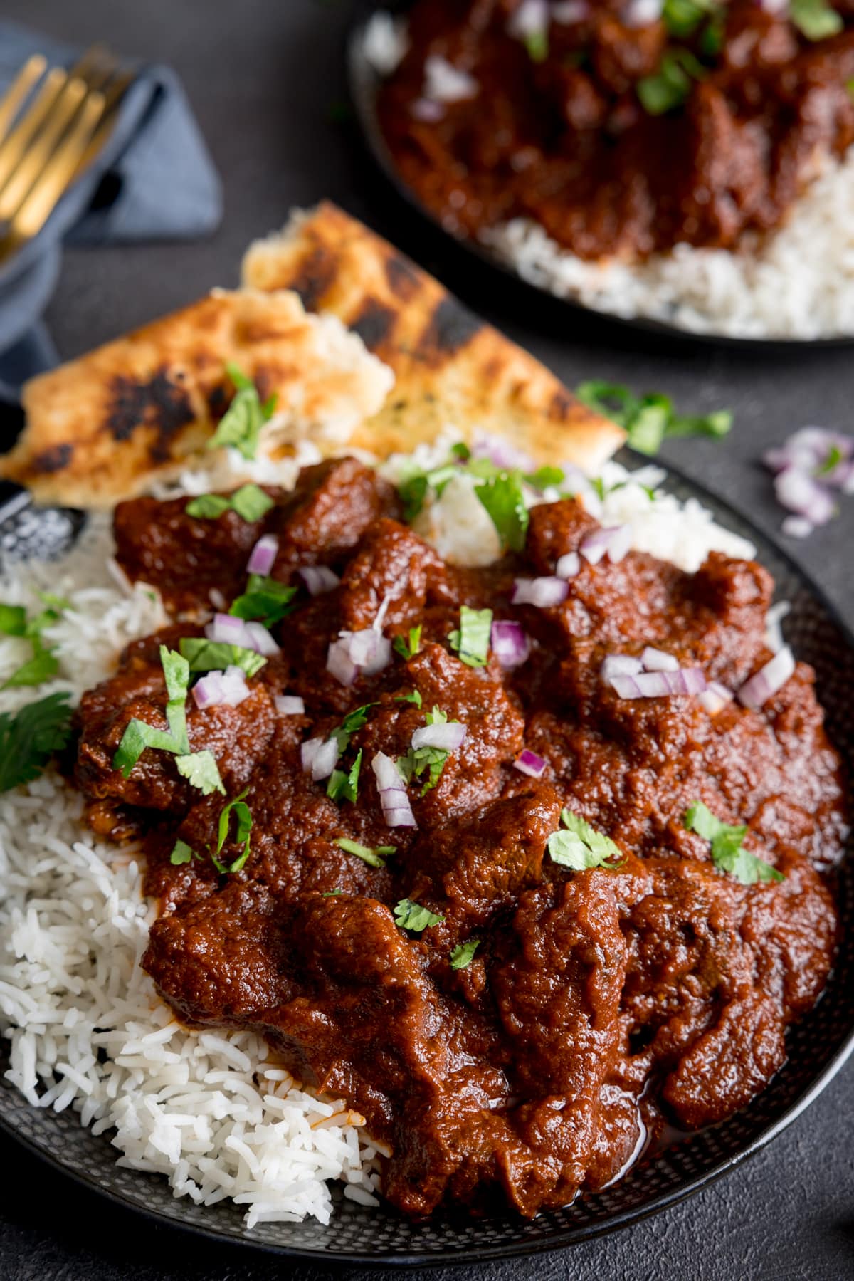 Close up image of slow cooked beef madras in a black bowl with rice and pieces of naan bread. There is coriander (cilantro) sprinkled on top. The bowl is on a dark surface and there is a further bowl of madras in the background.