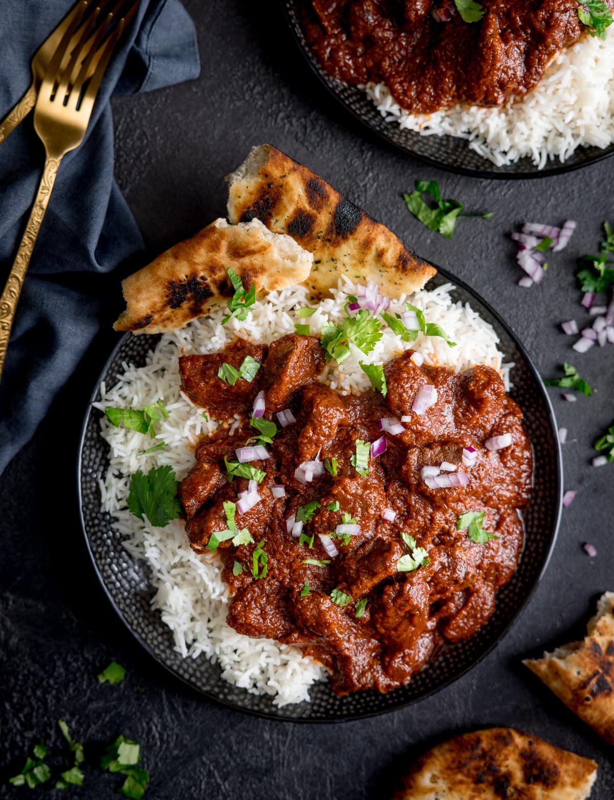 Tall overhead image of slow cooked beef madras in a black bowl with rice and pieces of naan bread. There is coriander (cilantro) sprinkled on top. The bowl is on a dark surface, next to a blue napkin, gold fork, more coriander and naan, and a further bowl of madras.