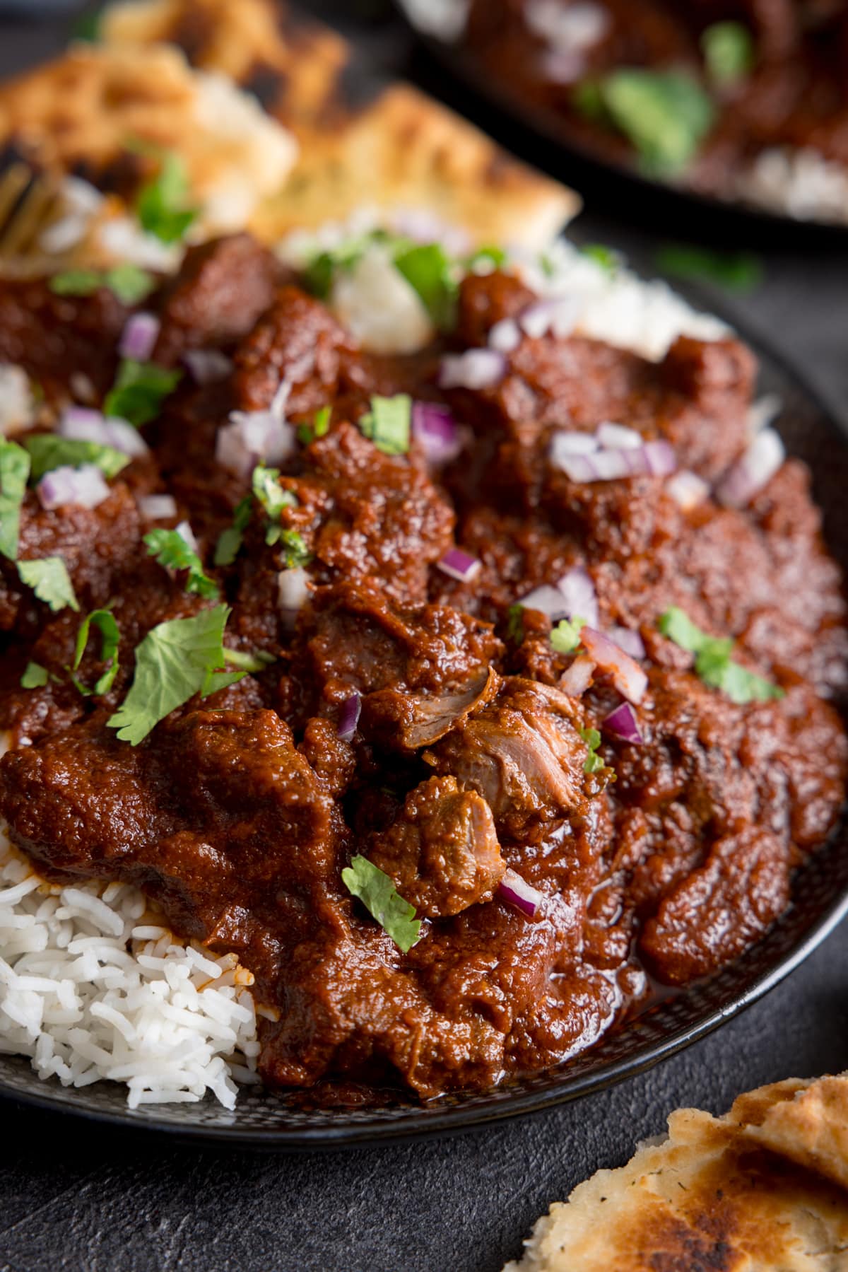 Close up image of slow cooked beef madras in a black bowl with rice and pieces of naan bread. There is coriander (cilantro) sprinkled on top. The bowl is on a dark surface.