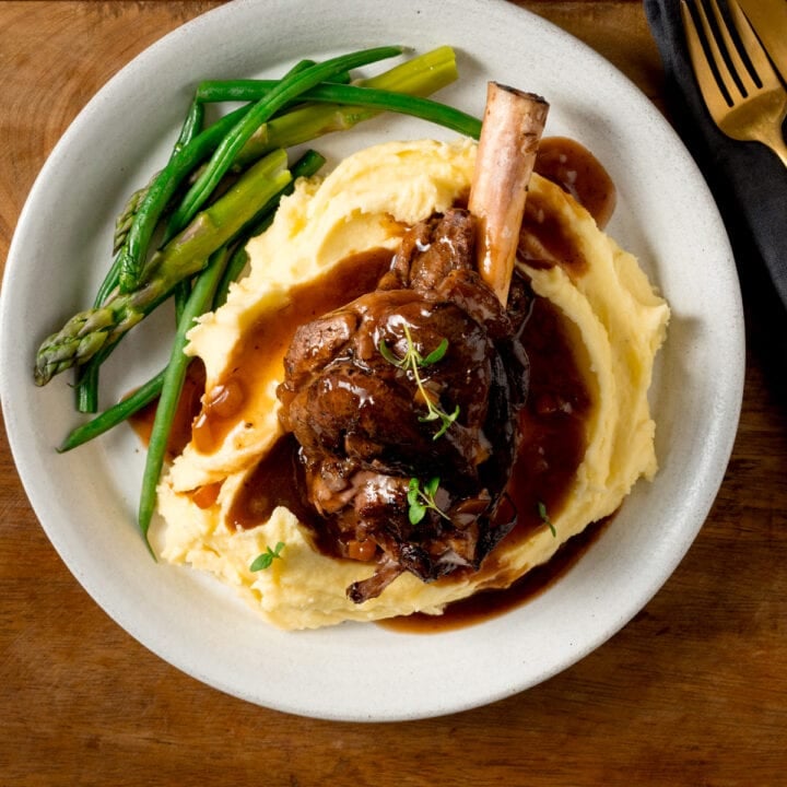 Square image of a slow cooked lamb shank on a pile of mashed potato with green beans and asparagus on a white plate. There is gravy and sprigs of fresh thyme on the lamb shank. The plate is on a wooden table next to a gold fork.