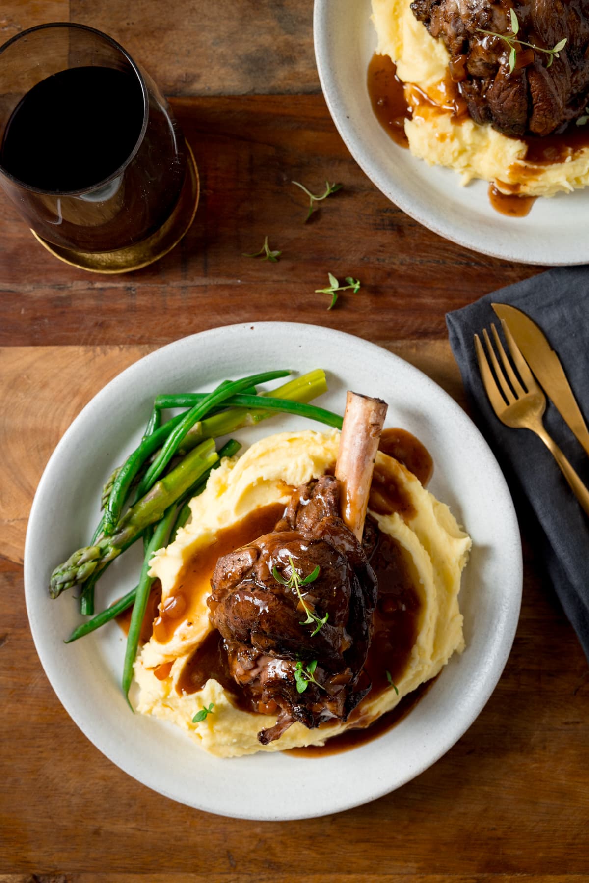Slow cooked lamb shank on a pile of mashed potato with green beans and asparagus on a white plate. There is gravy and sprigs of fresh thyme on the lamb shank. The plate is on a wooden table. There is a further plate with lamb shank and mashed potato at the top of the frame, along with a glass of red wine and a gold knife and fork on a grey napkin.