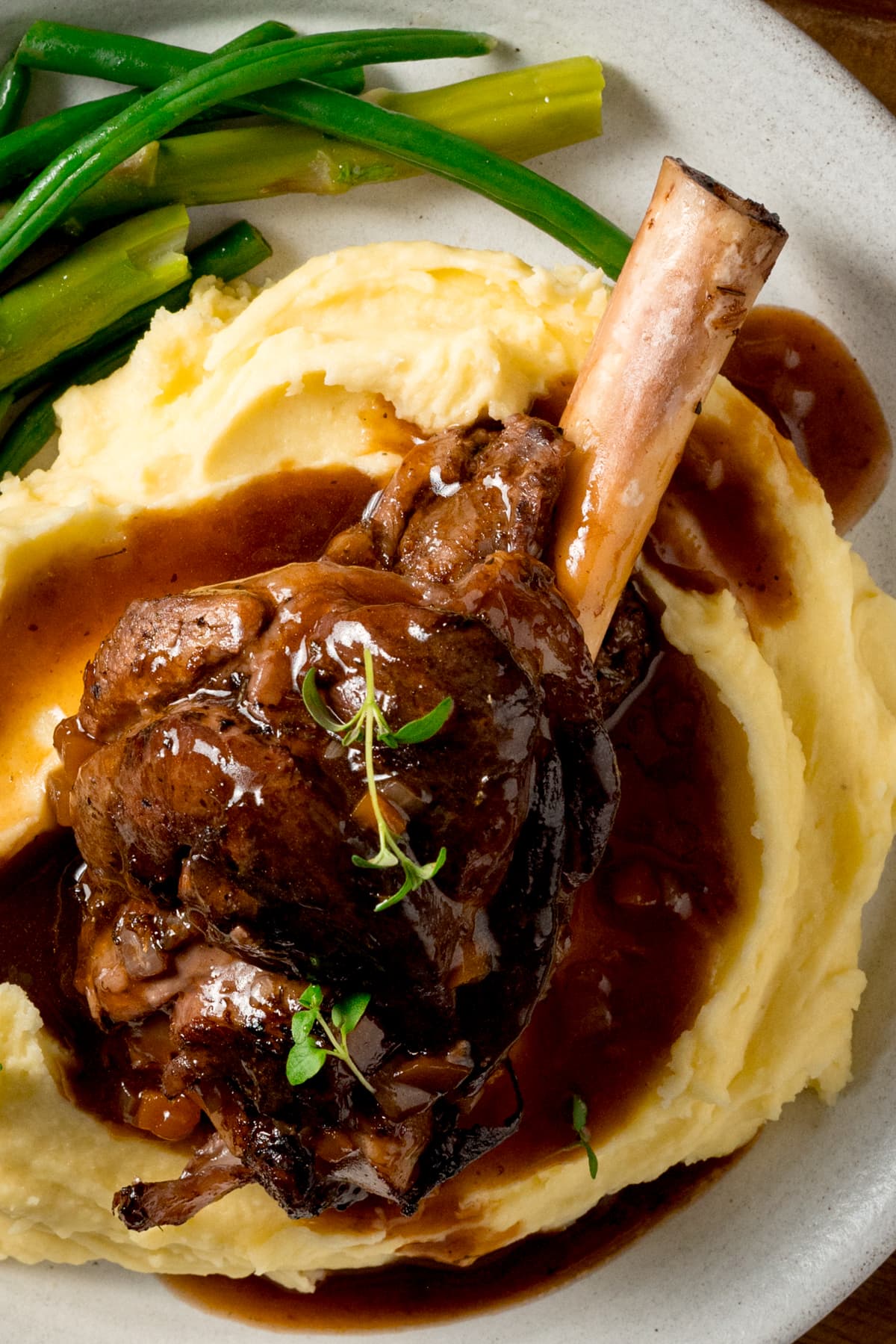 Slow cooked lamb shank on a pile of mashed potato with green beans and asparagus on a white plate. There is gravy and sprigs of fresh thyme on the lamb shank.