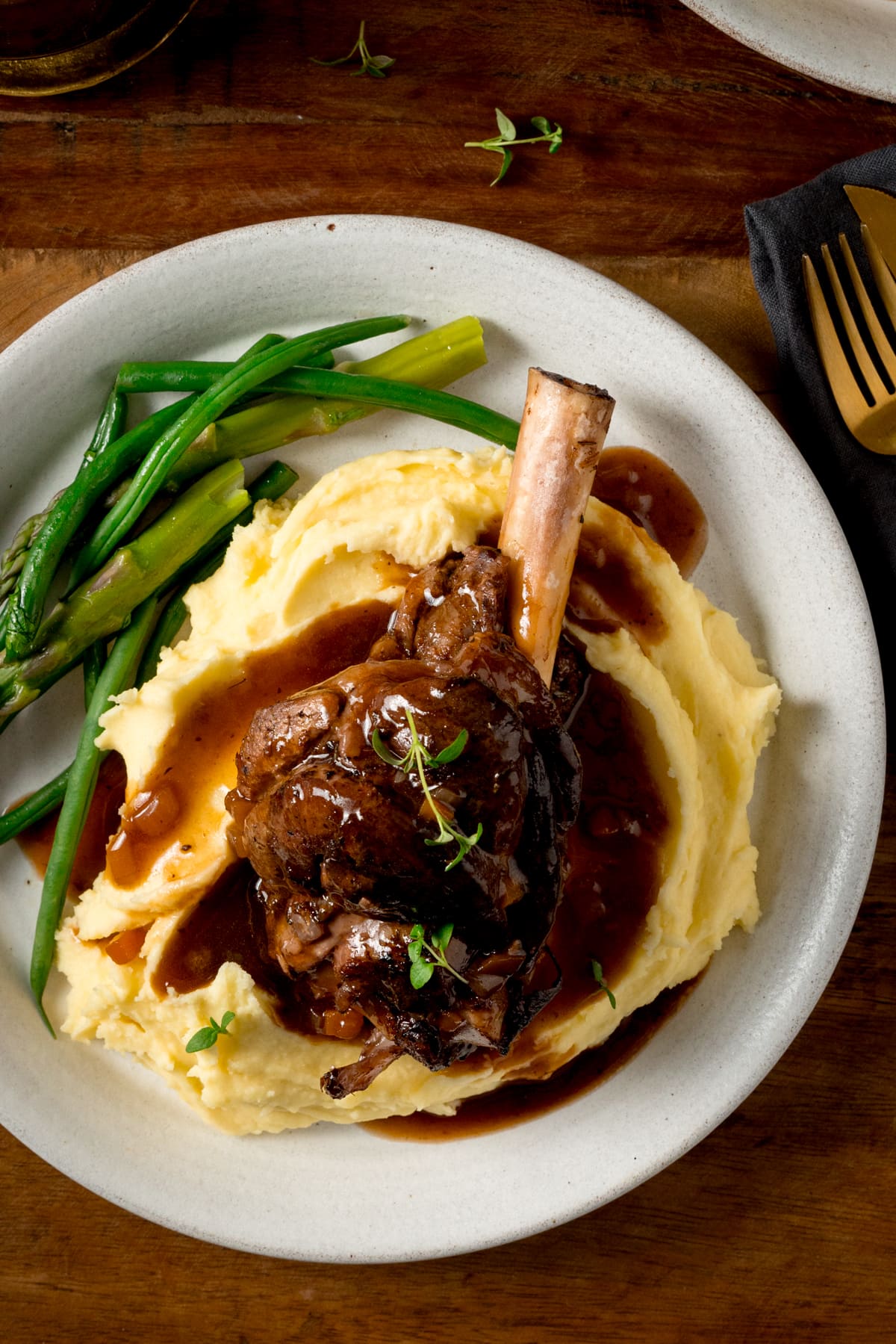 Slow cooked lamb shank on a pile of mashed potato with green beans and asparagus on a white plate. There is gravy and sprigs of fresh thyme on the lamb shank. The plate is on a wooden table.