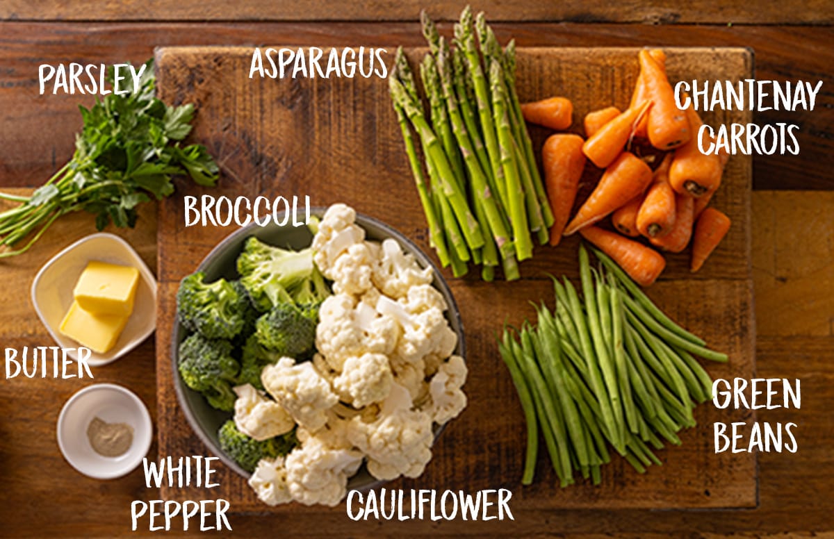 Ingredients for steamed vegetable medley on a wooden board. This includes carrots, cauliflower, broccoli, green beans, asparagus, butter and white pepper. The ingredients have a text overlay showing what each ingredient is.