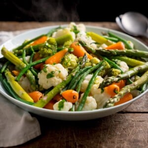 A square side-on image of a large light blue serving bowl, filled with mixed steamed vegetables. The bowl is on a wooden table. There is steam rising from the bowl. There is a white napkin and silver serving spoon next to the bowl.