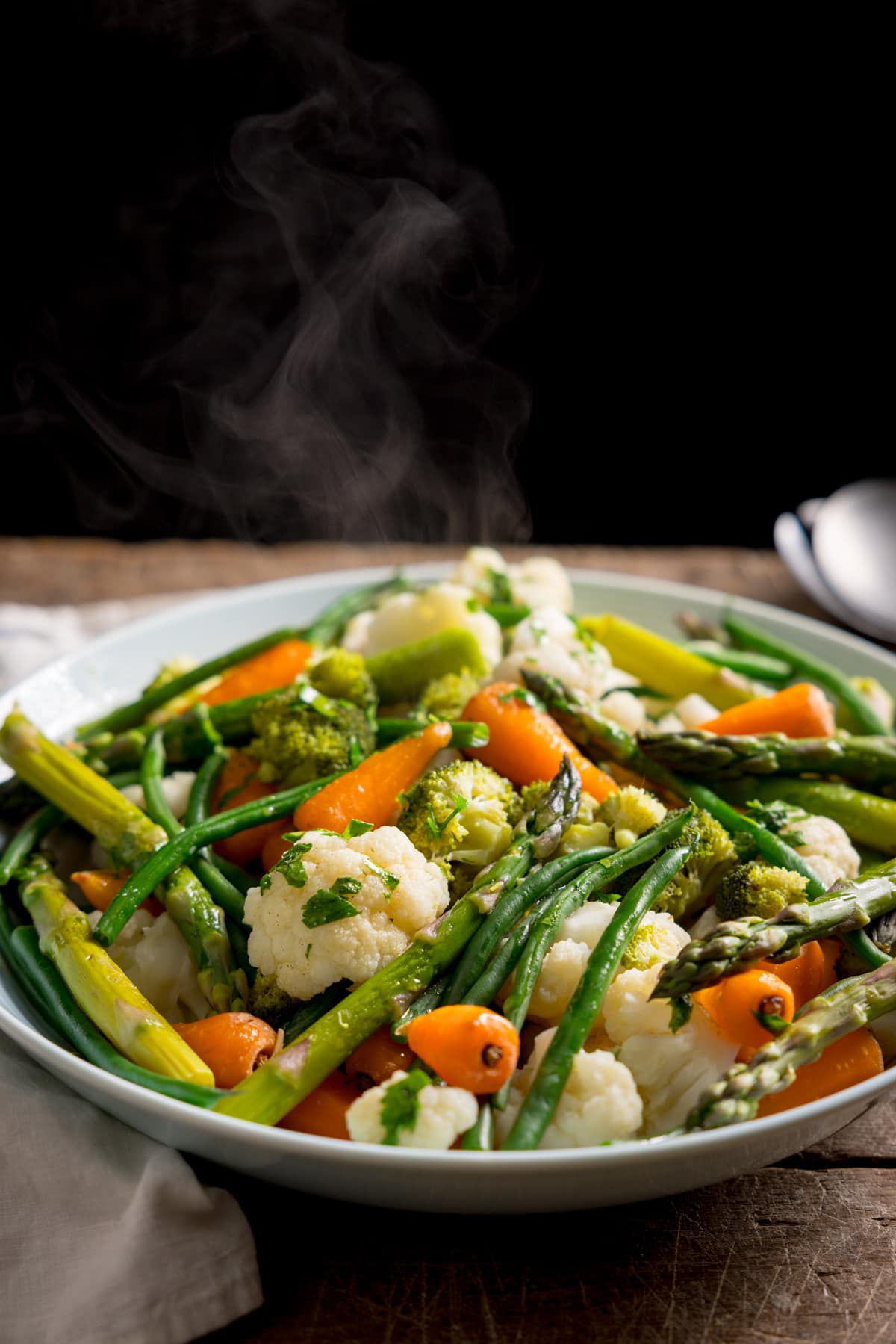 A tall side-on image of a large light blue serving bowl, filled with mixed steamed vegetables. The bowl is on a wooden table. There is steam rising from the bowl.