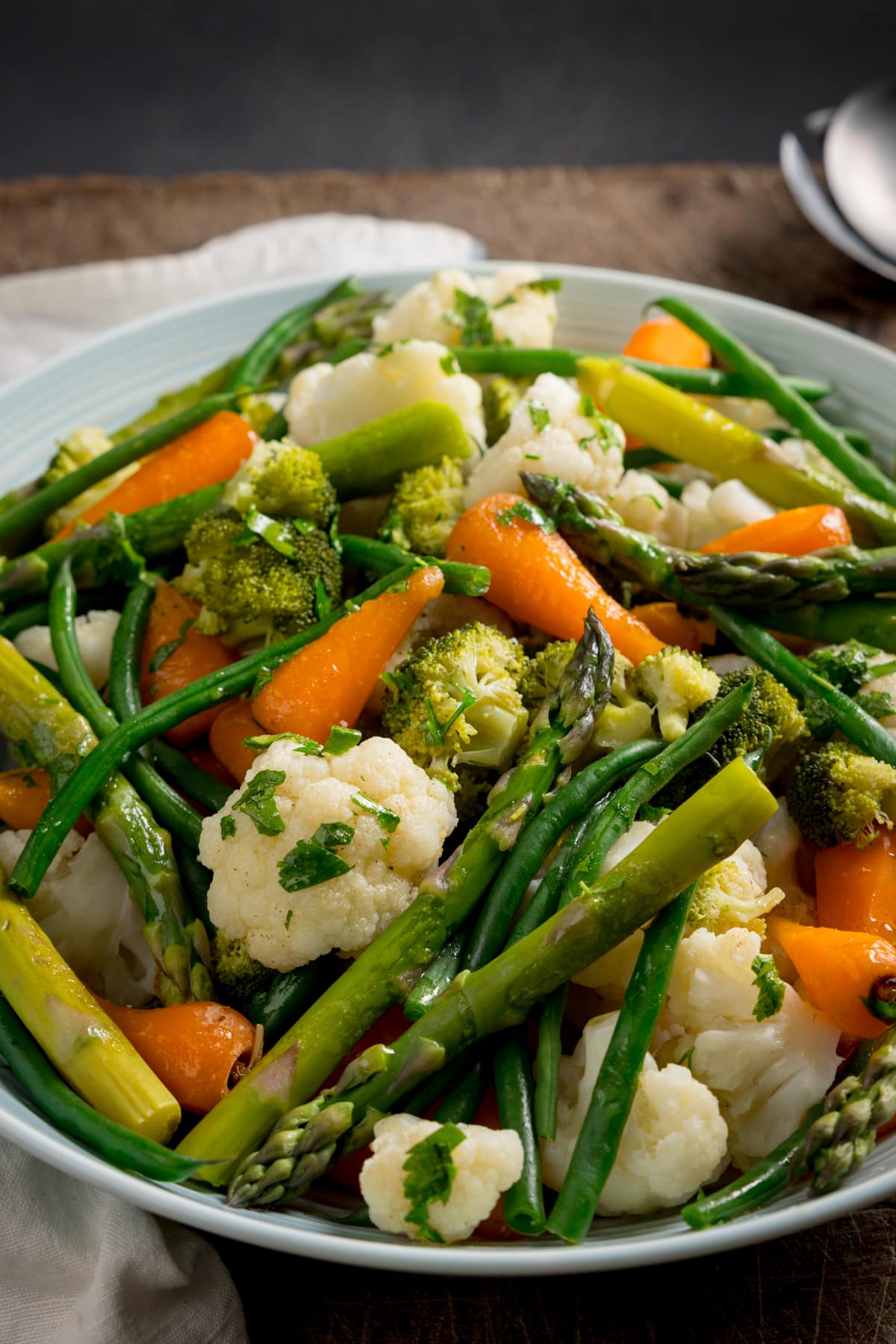 A large light blue serving bowl, filled with mixed steamed vegetables. The bowl is on a wooden table next to a white napkin and silver serving spoon.