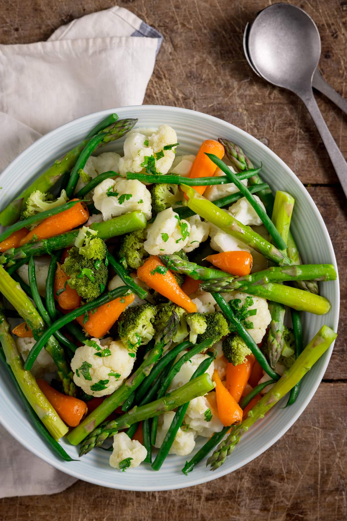 Overhead image of a large light blue serving bowl, filled with mixed steamed vegetables. The bowl is on a wooden table next to a white napkin and silver serving spoon.