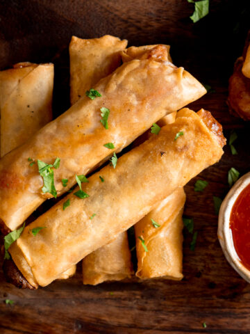 Overhead picture of a stack of 5 cooked vegetable spring rolls on a wooden board with a small pot of sweet chilli sauce.