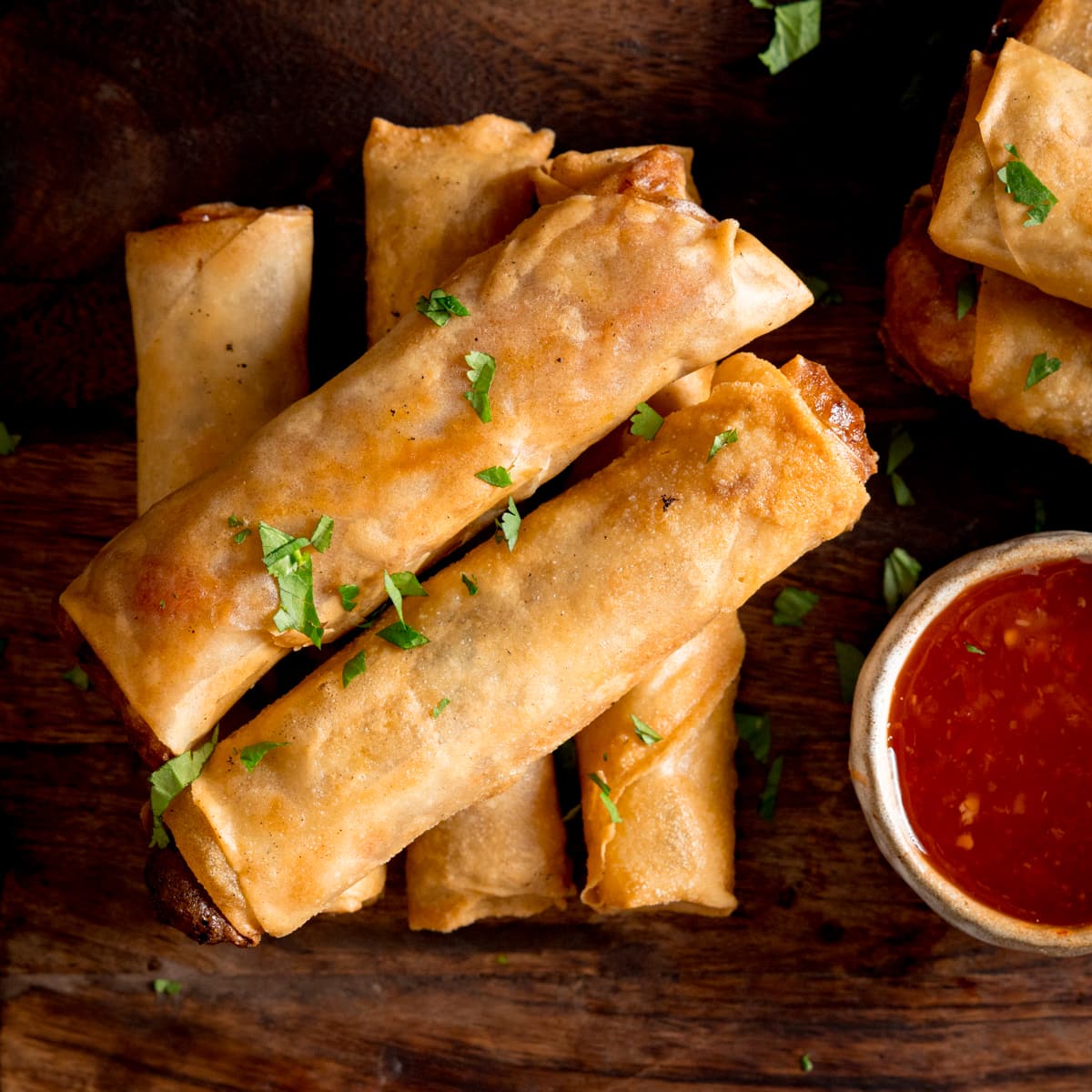 Overhead picture of a stack of 5 cooked vegetable spring rolls on a wooden board with a small pot of sweet chilli sauce.