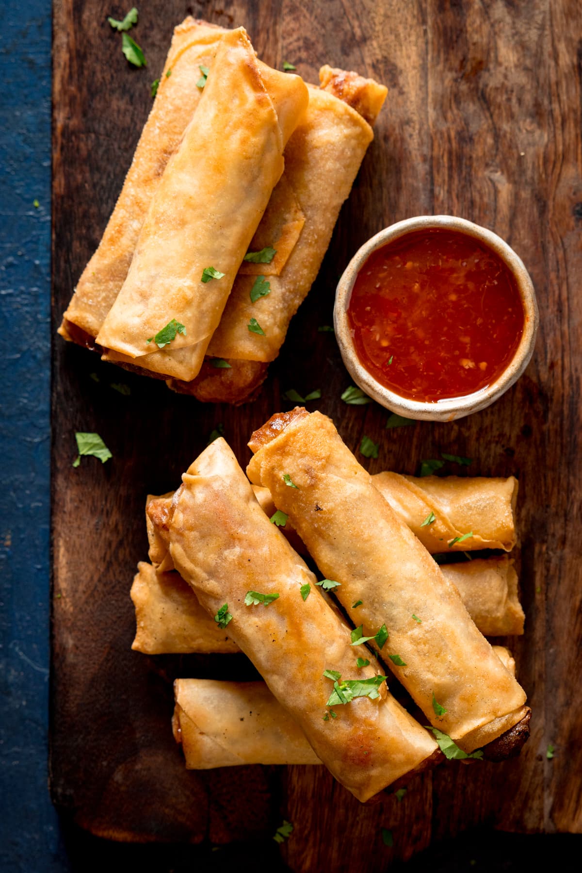 Overhead picture of 2 stacks of vegetable spring rolls on a wooden board with a small pot of sweet chilli sauce, all sprinkled with a little fresh coriander.