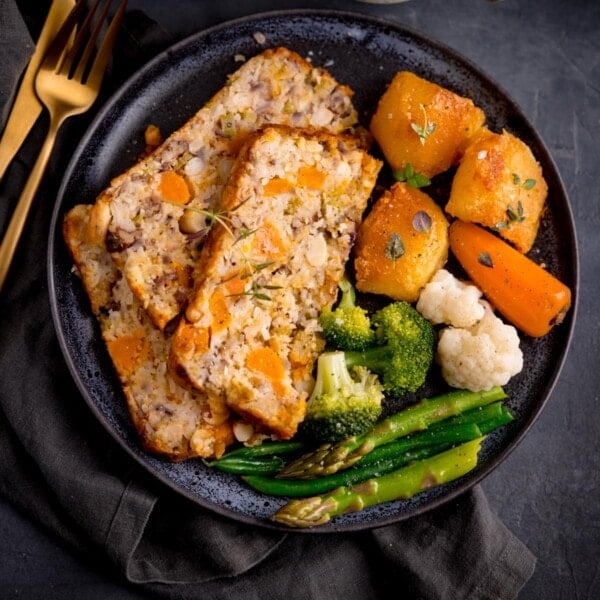 Three slices of vegetarian nut roast on a dark blue plate with vegetable and potatoes.